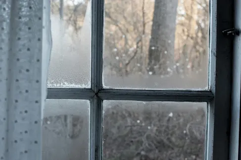 Condensation on a window pane, with a curtain to the left and trees visible outside